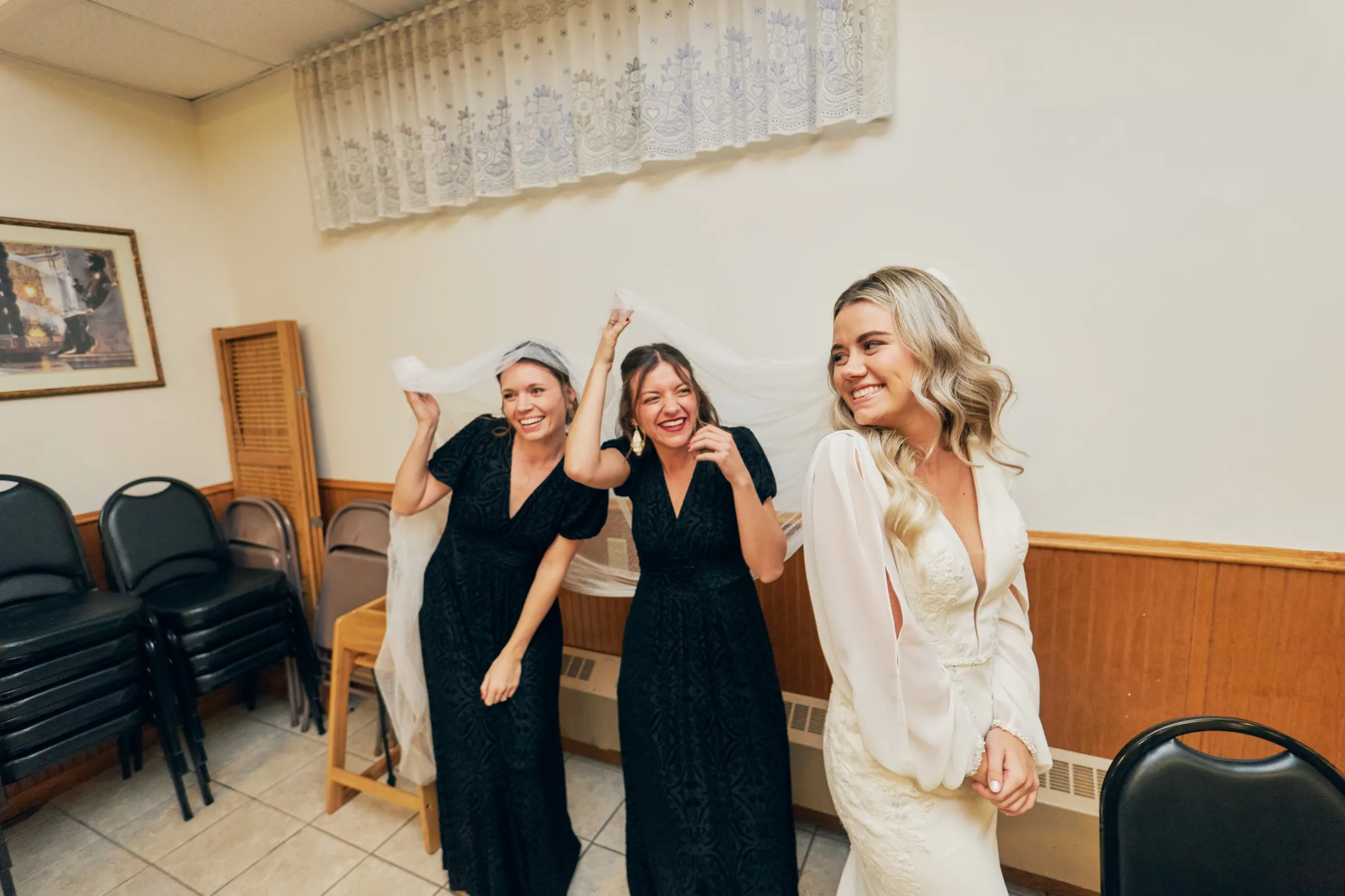 Two bridesmaids in black dresses laugh as they hold Madeline's long veil up above her in the getting-ready room — Tim Larsen Photography, Brainerd Lakes MN
