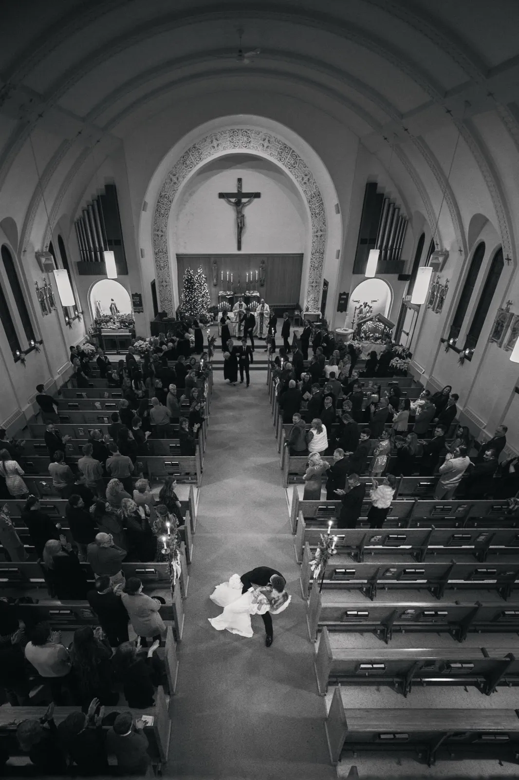 From a balcony view, the groom dips the bride in the center of a long church aisle as guests in the pews applaud beneath a vaulted nave and crucifix. — Tim Larsen Photography, Brainerd Lakes MN