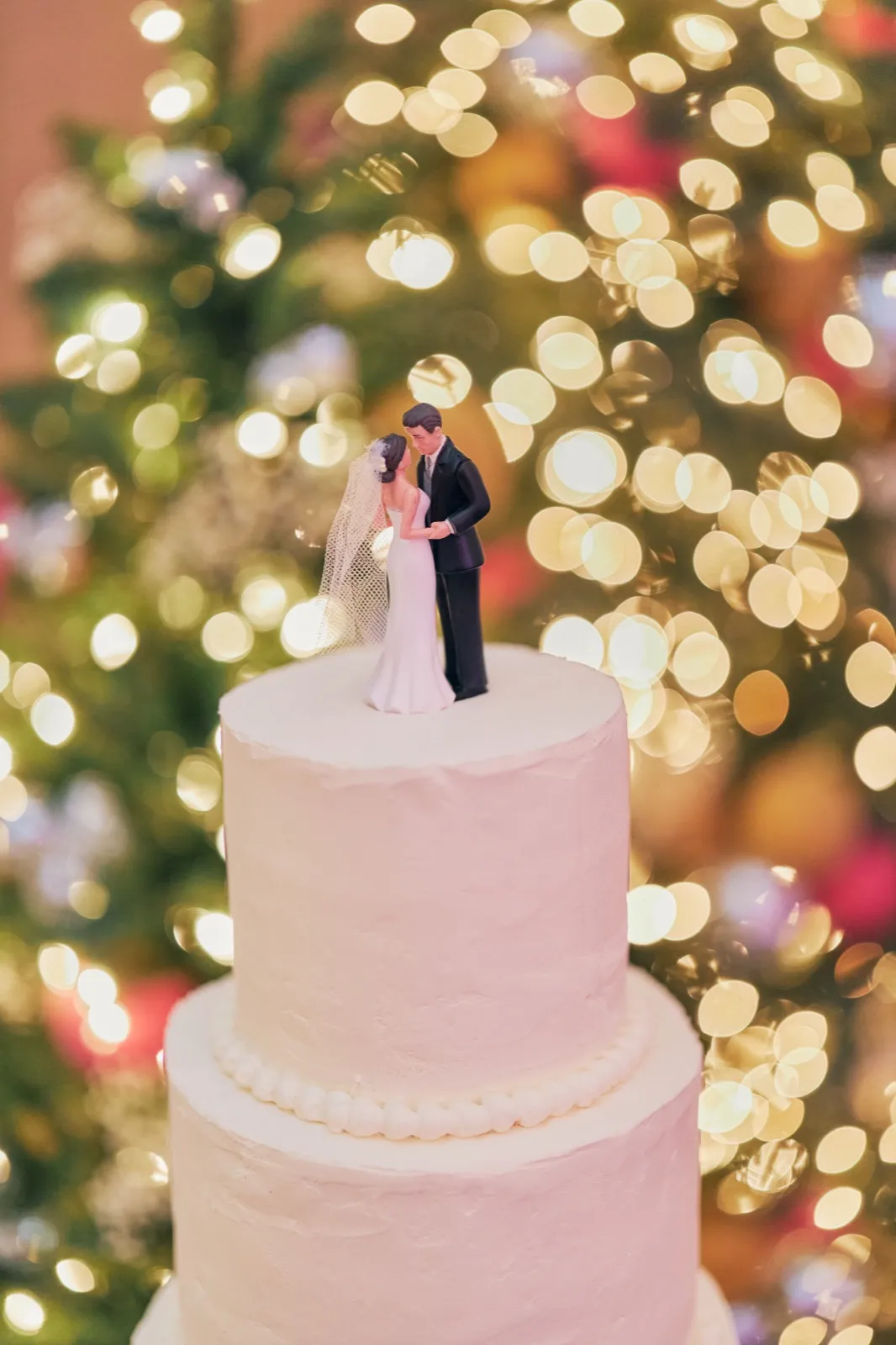 A white tiered wedding cake topped with a small bride-and-groom figurine stands in front of a brightly lit Christmas tree. — Tim Larsen Photography, Brainerd Lakes MN