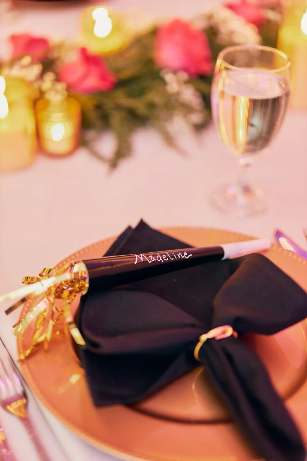 A handwritten 'Madeline' party popper rests on a black napkin atop a gold charger plate, with a champagne flute and pink rose garland softly blurred behind. — Tim Larsen Photography, Brainerd Lakes MN