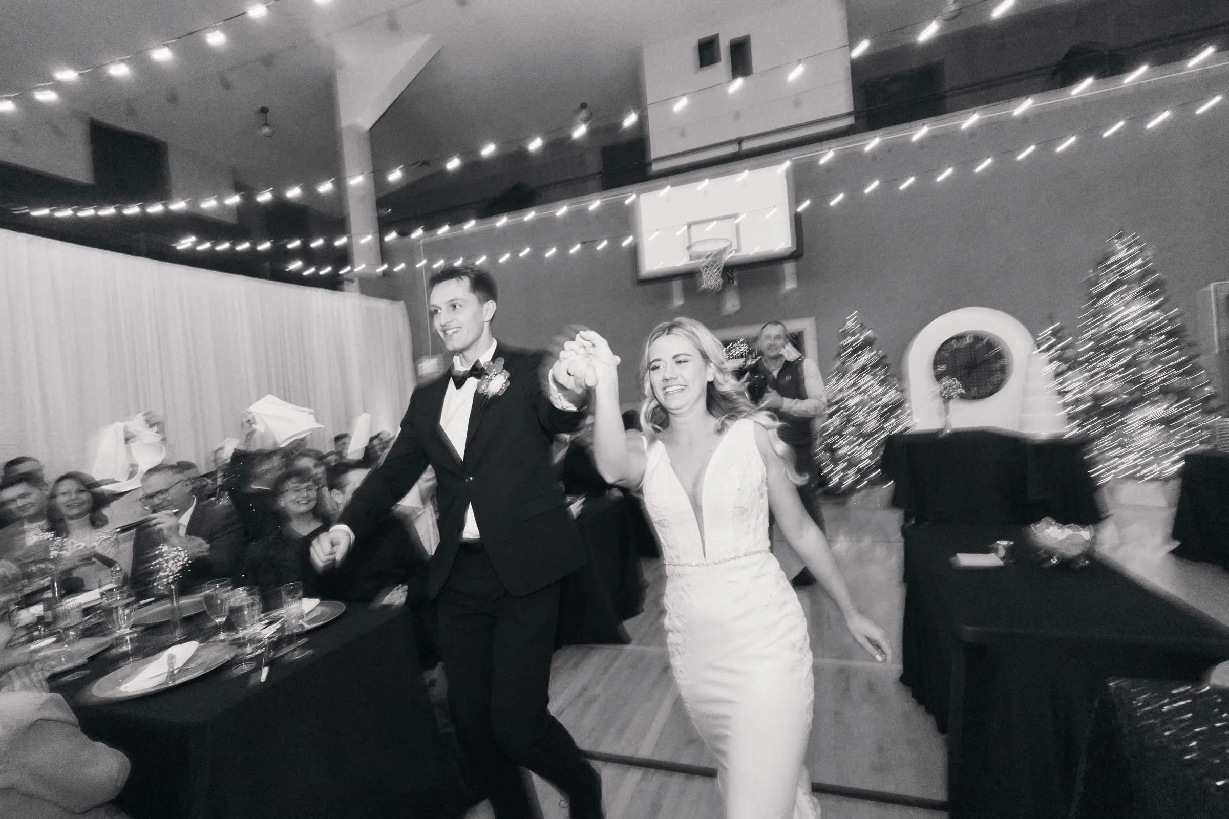Madeline and Sam enter their reception hand in hand with arms raised beneath a canopy of string lights, the gymnasium's basketball hoop and Christmas trees behind them — Tim Larsen Photography, Brainerd Lakes MN