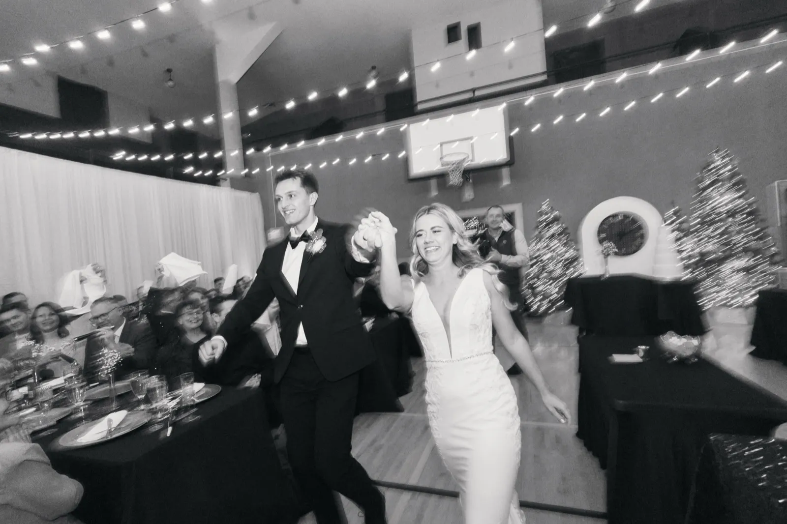 The bride and groom enter their reception hand-in-hand with arms raised, walking past seated guests beneath a canopy of string lights with a basketball hoop and Christmas trees visible in the background. — Tim Larsen Photography, Brainerd Lakes MN
