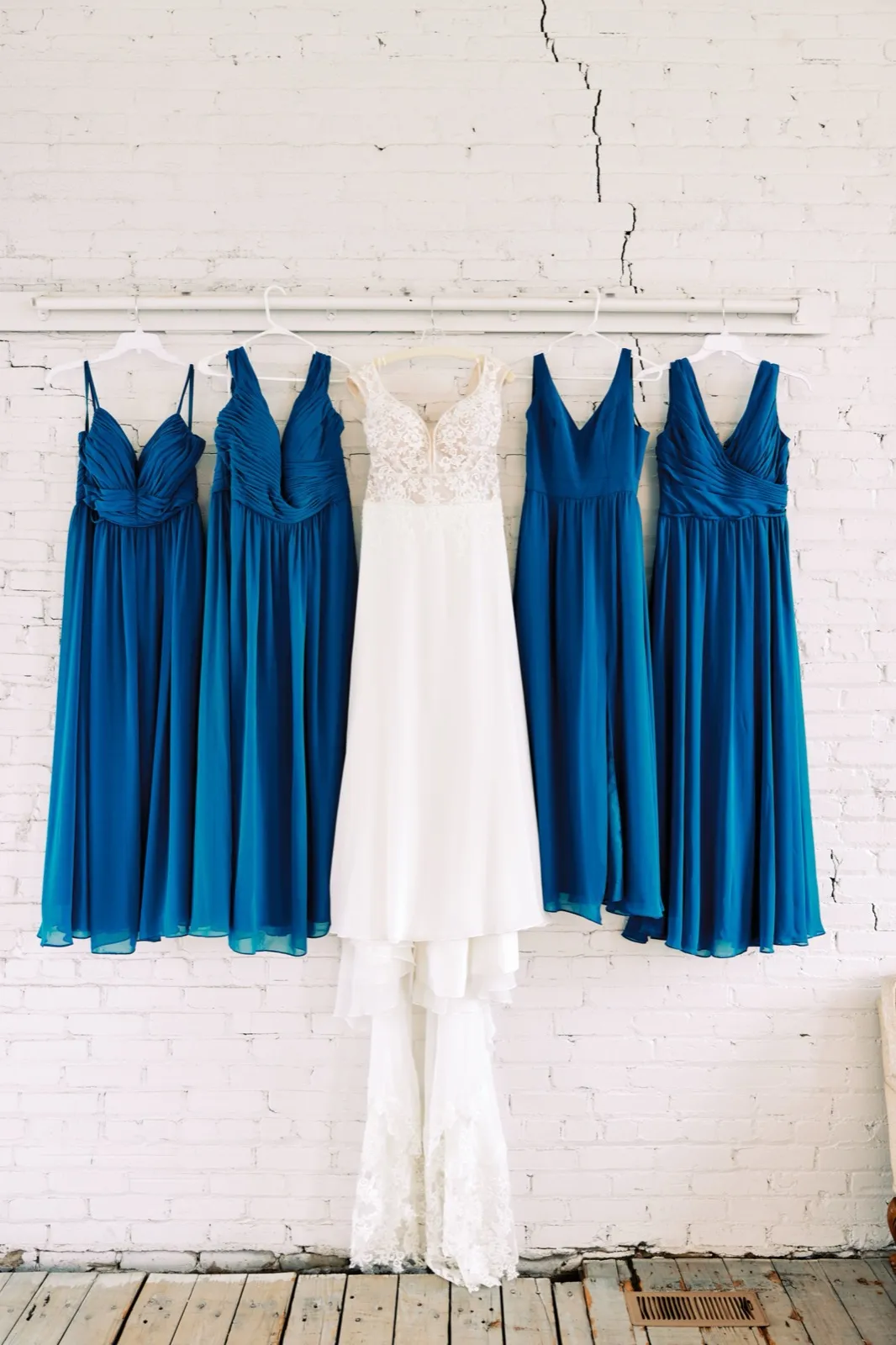 Maggie's white lace gown hangs centered between four blue bridesmaid dresses on a white-painted exposed brick wall at Northern Pacific Center — Tim Larsen Photography, Brainerd Lakes MN