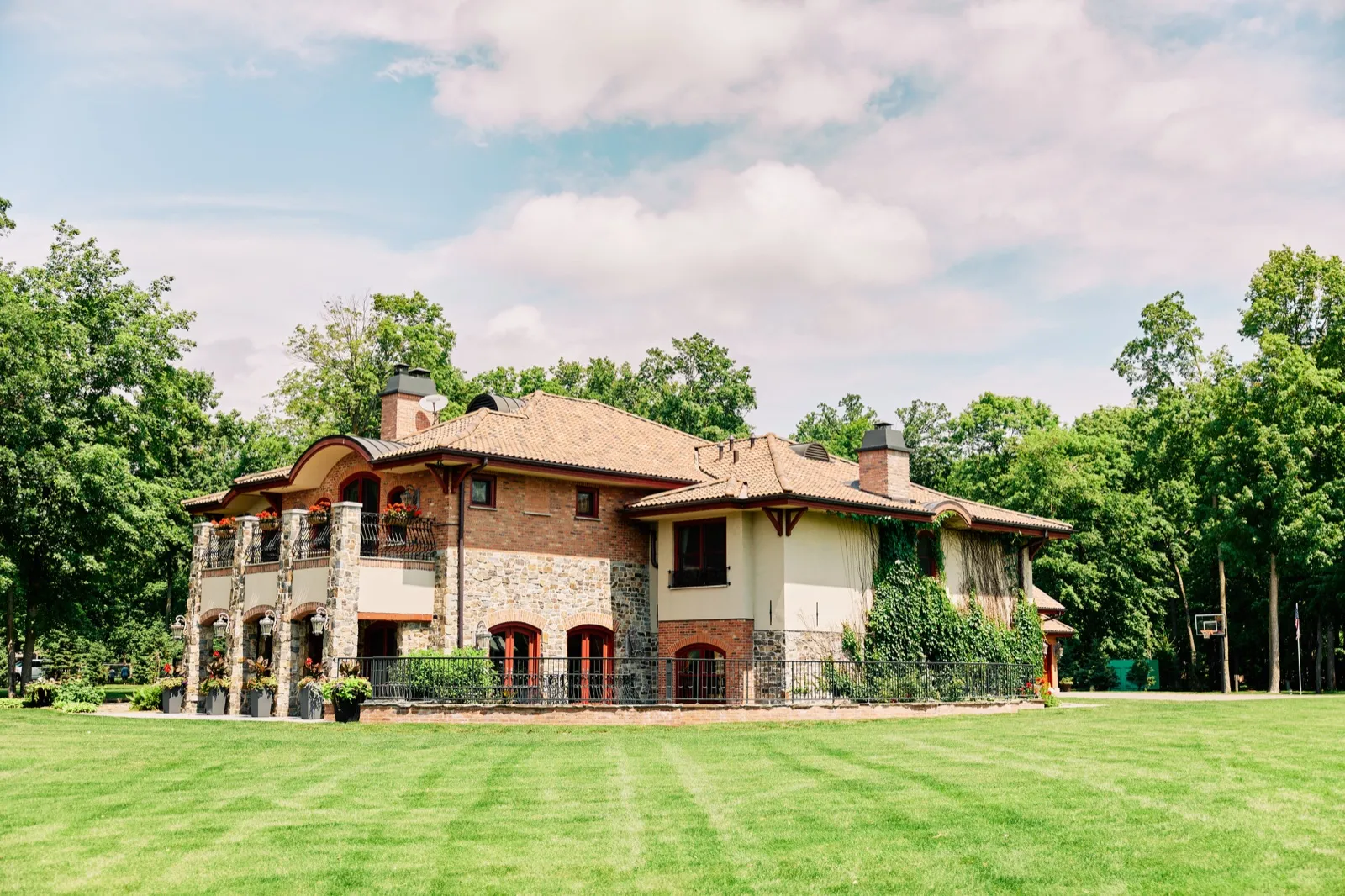 A large stone and brick lake home with arched balconies and ivy-covered walls sits beyond a wide green lawn under a partly cloudy summer sky. — Tim Larsen Photography, Brainerd Lakes MN