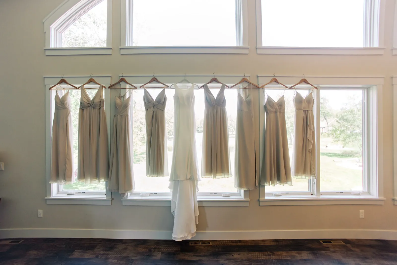 A wedding gown hangs centered among nine neutral-toned bridesmaid dresses, all backlit by a tall wall of windows in a bridal suite. — Tim Larsen Photography, Brainerd Lakes MN