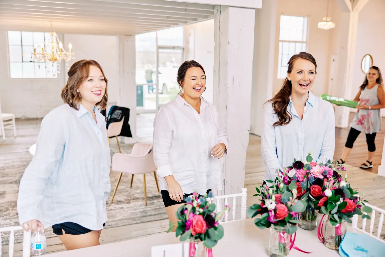 Maggie laughs with two bridesmaids in white button-down shirts beside their bouquets in the bright getting-ready room — Tim Larsen Photography, Brainerd Lakes MN