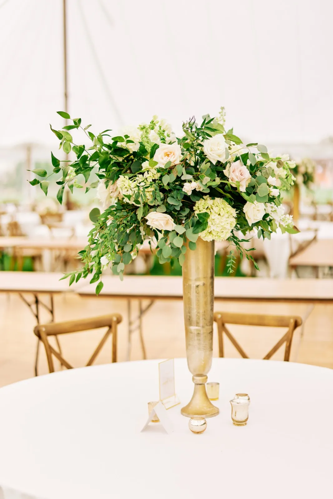 A tall gold vase holds a lush arrangement of white roses, hydrangeas, and trailing greenery on a sweetheart table inside a sailcloth tent. — Tim Larsen Photography, Brainerd Lakes MN