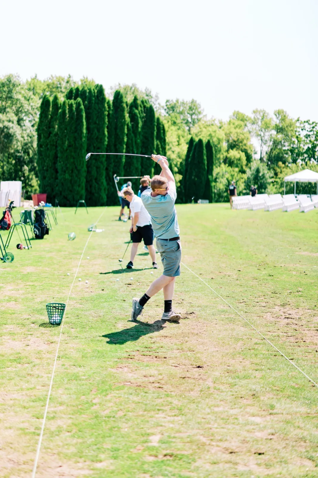 A guest in shorts and a t-shirt swings a golf club at the top of his backswing on a sunny driving range, with other guests hitting balls behind him. — Tim Larsen Photography, Twin Cities MN