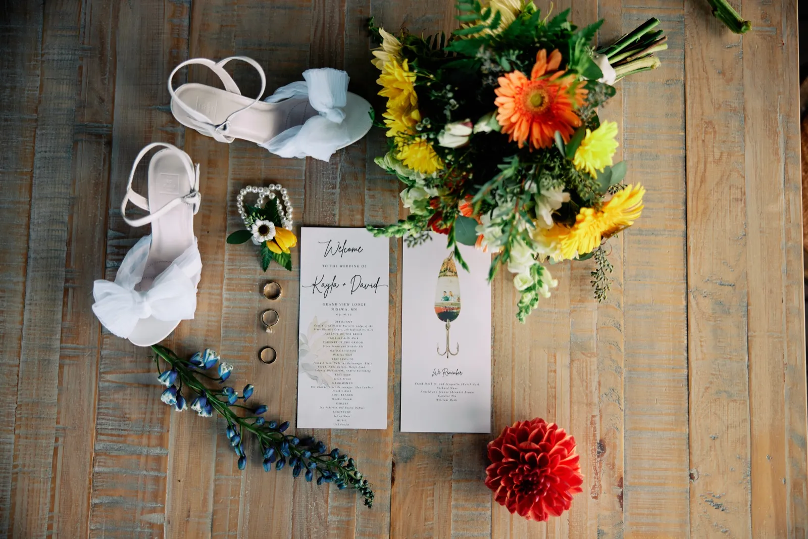 An overhead flat-lay on a weathered wood floor showing the bride's white shoes, a vibrant sunflower and gerbera bouquet, the wedding program for Kayla and David at Grand View Lodge, two rings, a fishing-lure boutonniere, and sprigs of blue delphinium. — Tim Larsen Photography, Brainerd Lakes MN