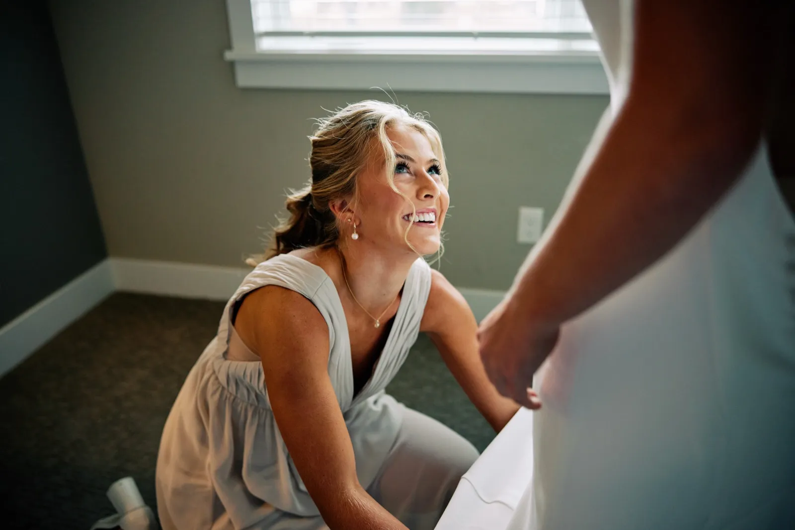 A bridesmaid kneels and looks up laughing at the bride while helping her into her wedding dress in a window-lit getting-ready room. — Tim Larsen Photography, Brainerd Lakes MN