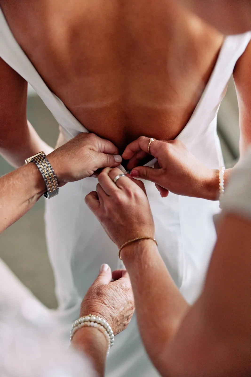 Multiple women's hands, including an elderly relative's, work together to fasten the back of the bride's open-back wedding dress. — Tim Larsen Photography, Brainerd Lakes MN