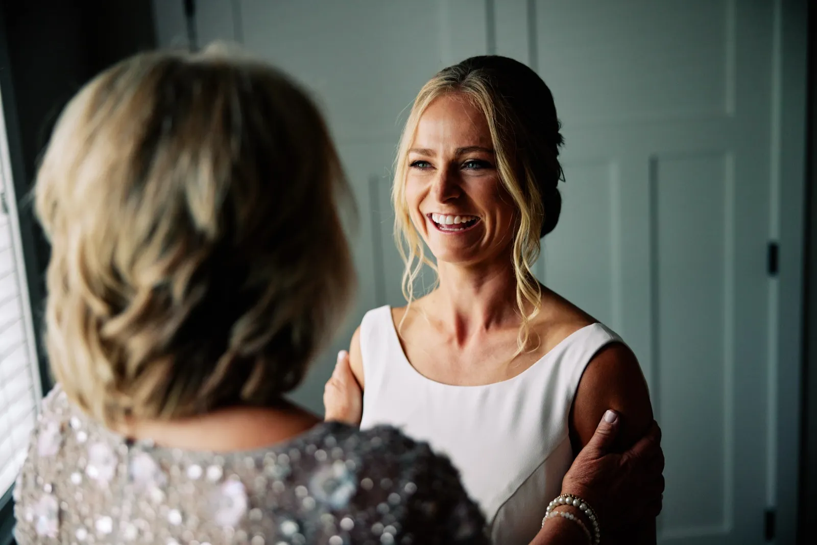 The bride laughs in window light as her mother, seen from behind, rests a hand on her shoulder in the bridal suite. — Tim Larsen Photography, Brainerd Lakes MN