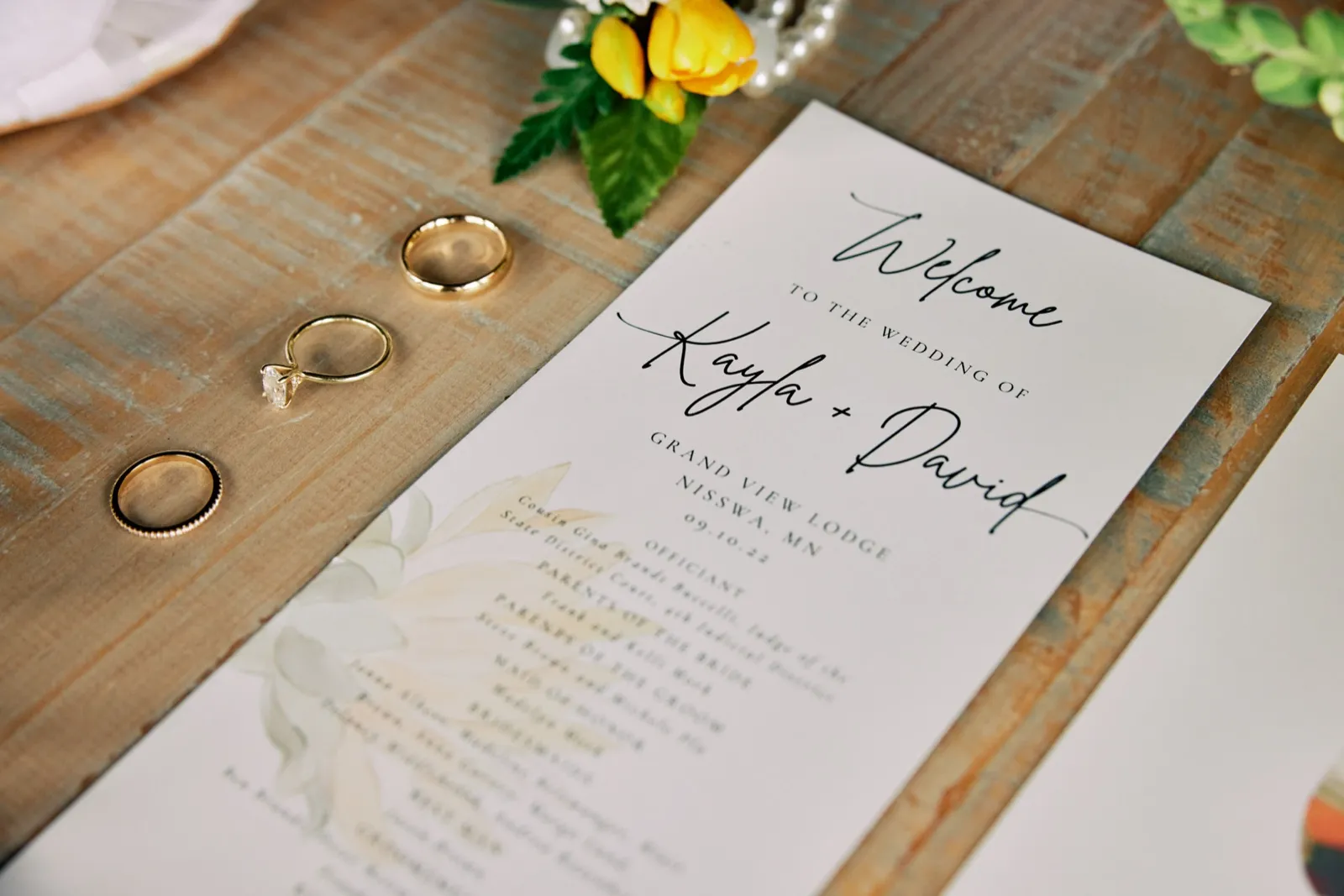 Two gold wedding bands and a solitaire engagement ring rest on a weathered wood surface beside a ceremony program reading 'Welcome to the wedding of Kayla + David, Grand View Lodge, Nisswa, MN, 09.10.22' with yellow tulips above. — Tim Larsen Photography, Brainerd Lakes MN