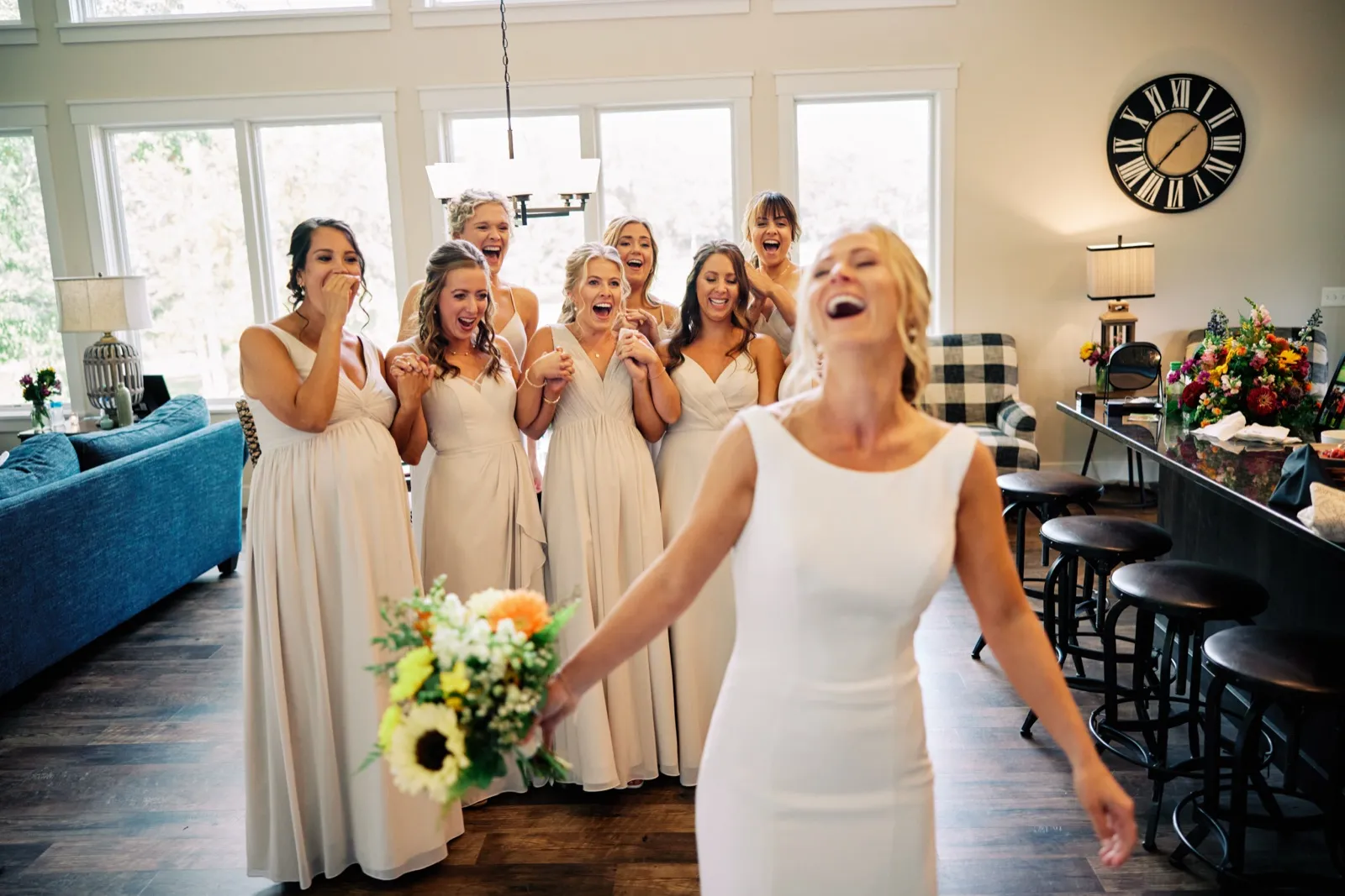 The bride laughs in the foreground while her bridesmaids react with open-mouthed delight behind her in a window-lit room. — Tim Larsen Photography, Brainerd Lakes MN