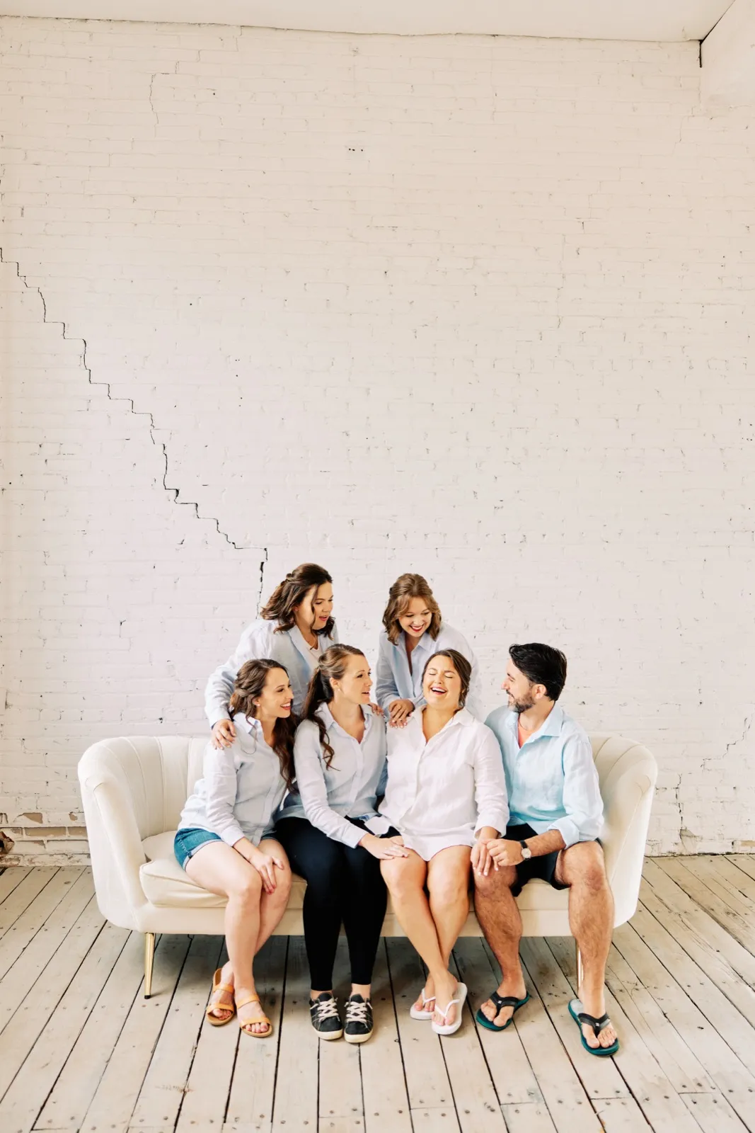 Maggie laughs on a cream sofa surrounded by her wedding party in button-down shirts against the tall white brick of Le Lier — Tim Larsen Photography, Brainerd Lakes MN