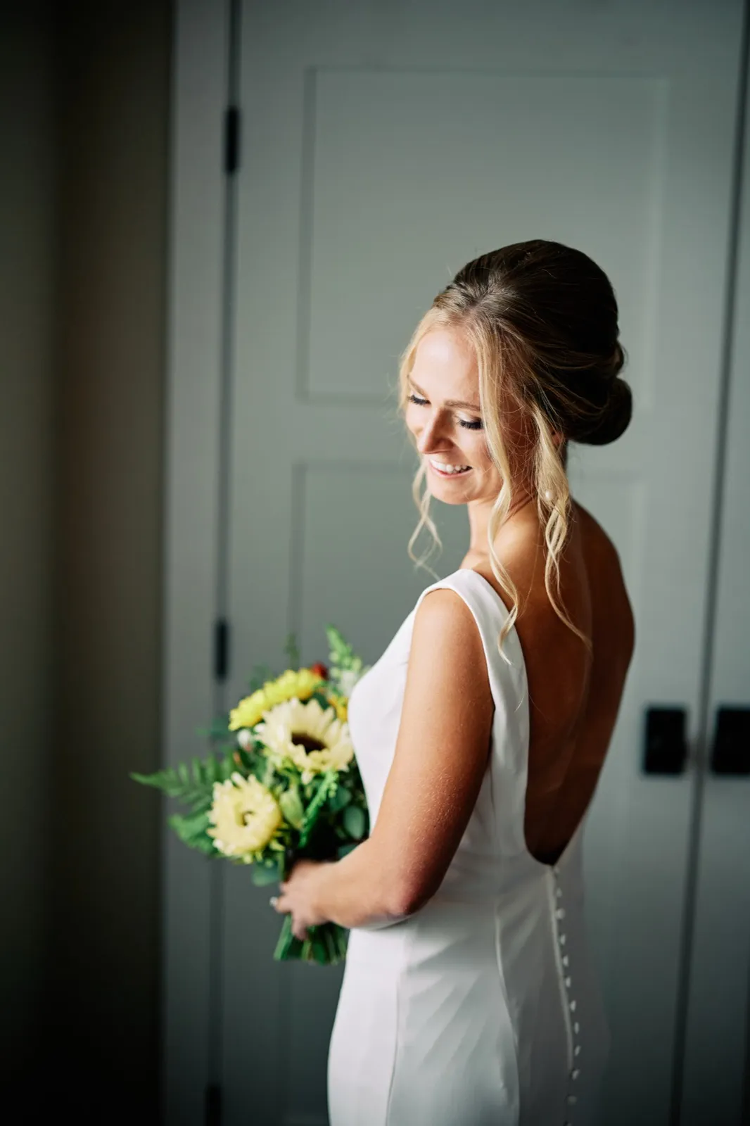 The bride stands in profile in a bridal suite, smiling down at her sunflower bouquet as window light catches her low bun and the open back of her white gown. — Tim Larsen Photography, Brainerd Lakes MN