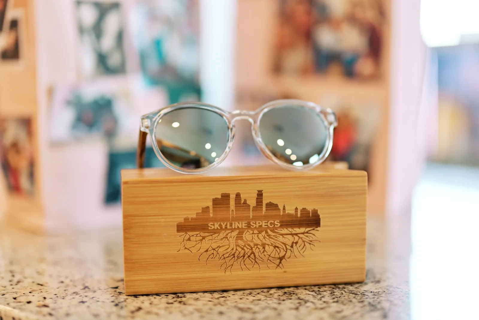 A pair of clear-framed mirrored sunglasses rests atop a bamboo case engraved with a city skyline and the words 'Skyline Specs.' — Tim Larsen Photography, Twin Cities MN