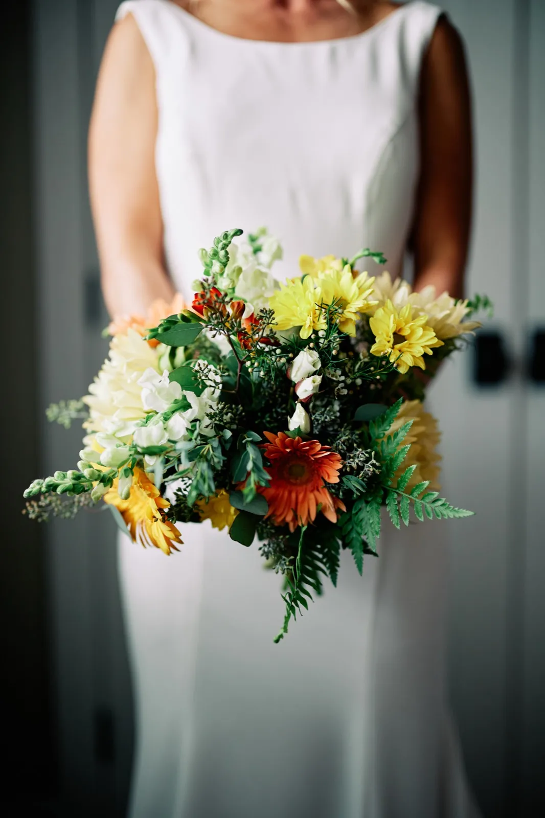 The bride holds an autumn bouquet of yellow chrysanthemums, orange gerbera daisies, white snapdragons, and ferns in front of her ivory dress. — Tim Larsen Photography, Brainerd Lakes MN