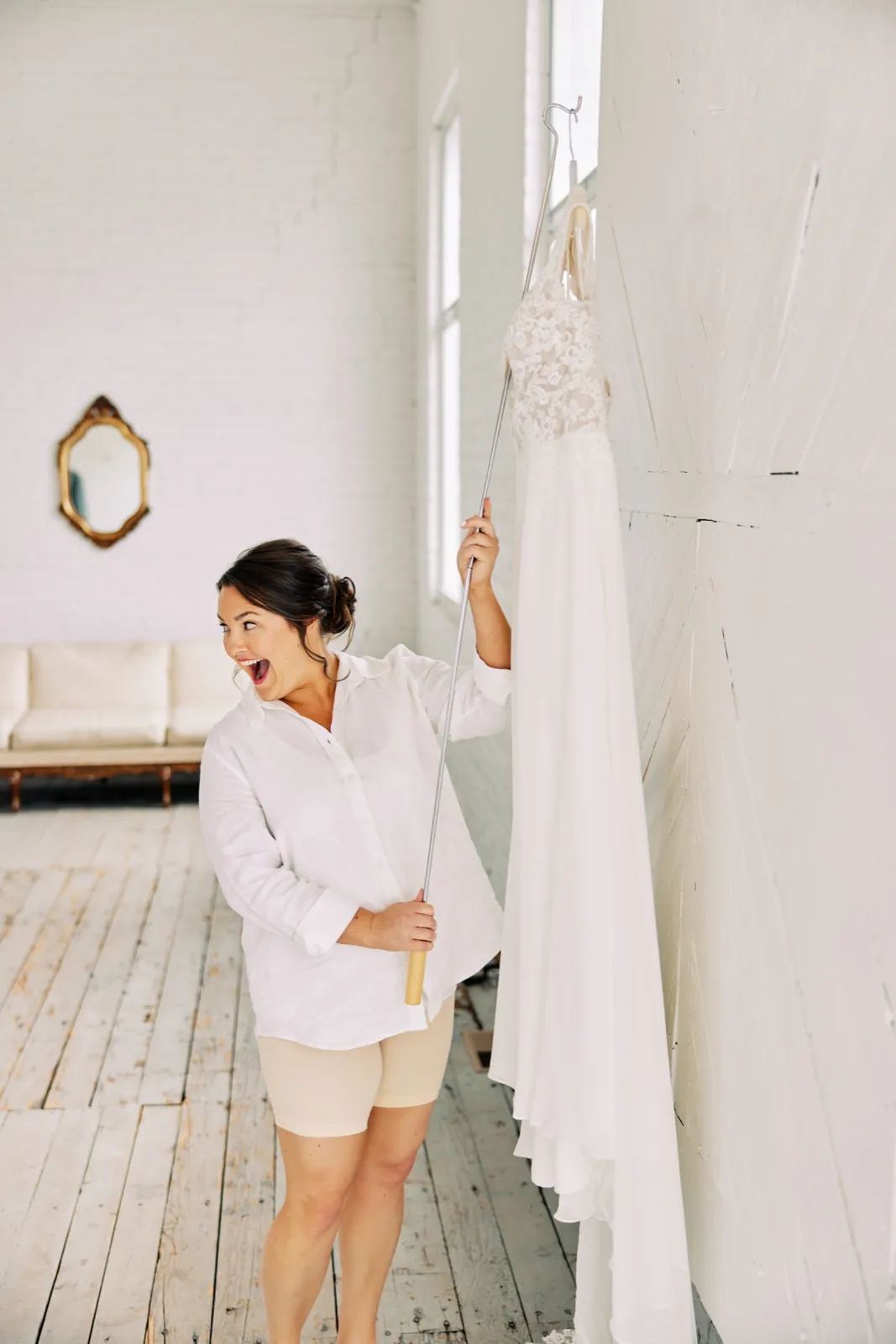 Maggie laughs as she uses a long hanger pole to reach her wedding dress high on the white brick wall of Le Lier — Tim Larsen Photography, Brainerd Lakes MN