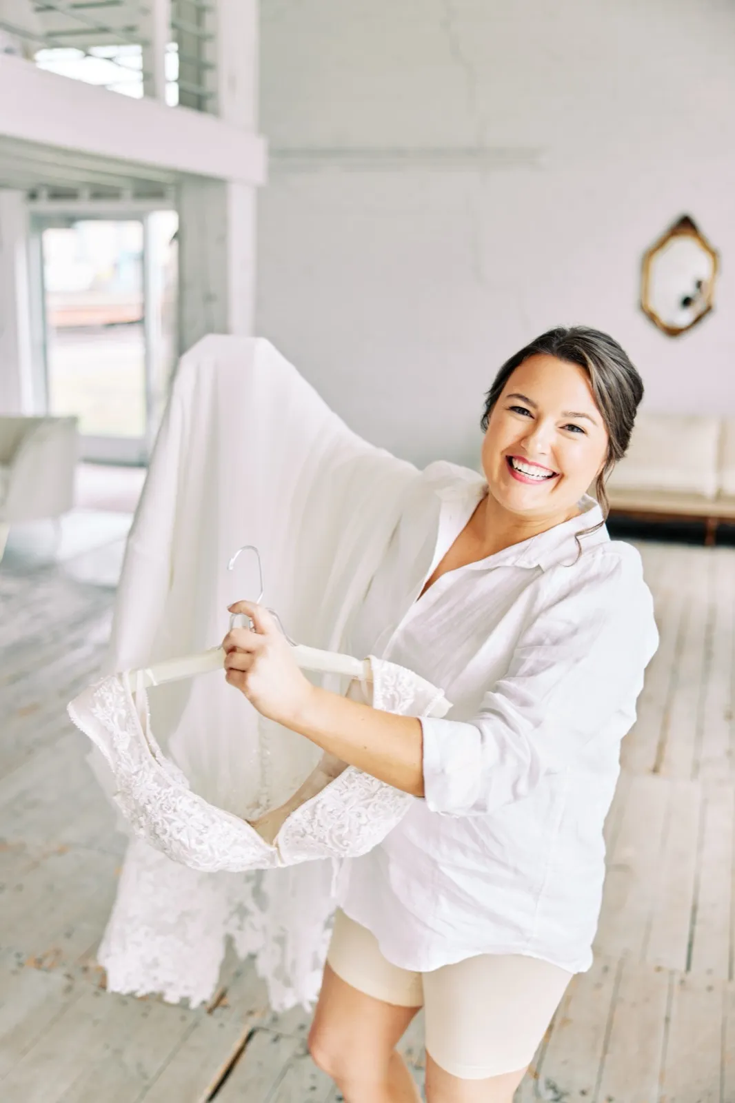 Maggie laughs while holding up her lace wedding dress on a hanger in a bright window-lit getting-ready room — Tim Larsen Photography, Brainerd Lakes MN