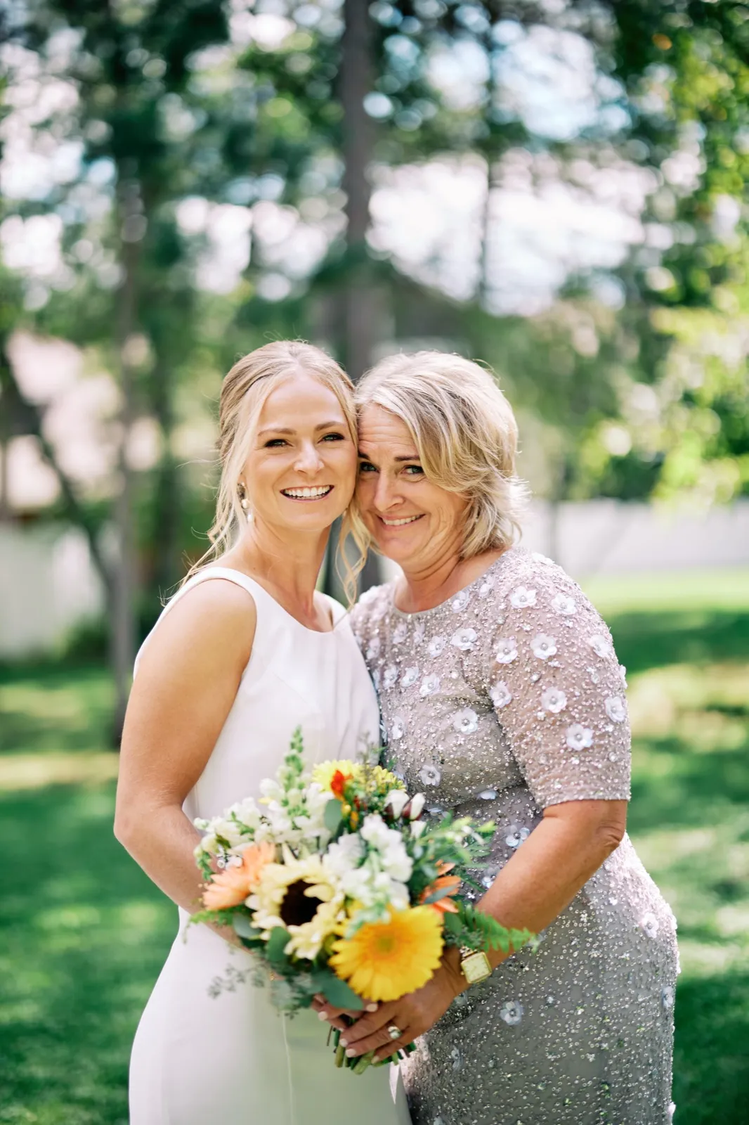 The bride and her mother stand cheek-to-cheek smiling at the camera, the bride holding a sunflower-and-rose bouquet on a sunlit lawn. — Tim Larsen Photography, Brainerd Lakes MN