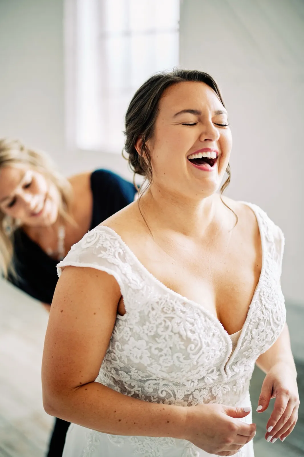 Maggie laughs with her head thrown back as a woman fastens the back of her lace gown in a window-lit room — Tim Larsen Photography, Brainerd Lakes MN