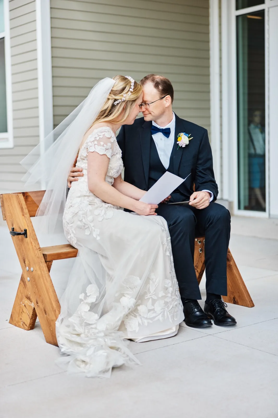 The bride and groom sit close on a wooden bench, foreheads pressed together, holding a vow card between them on a covered porch. — Tim Larsen Photography, Minnesota