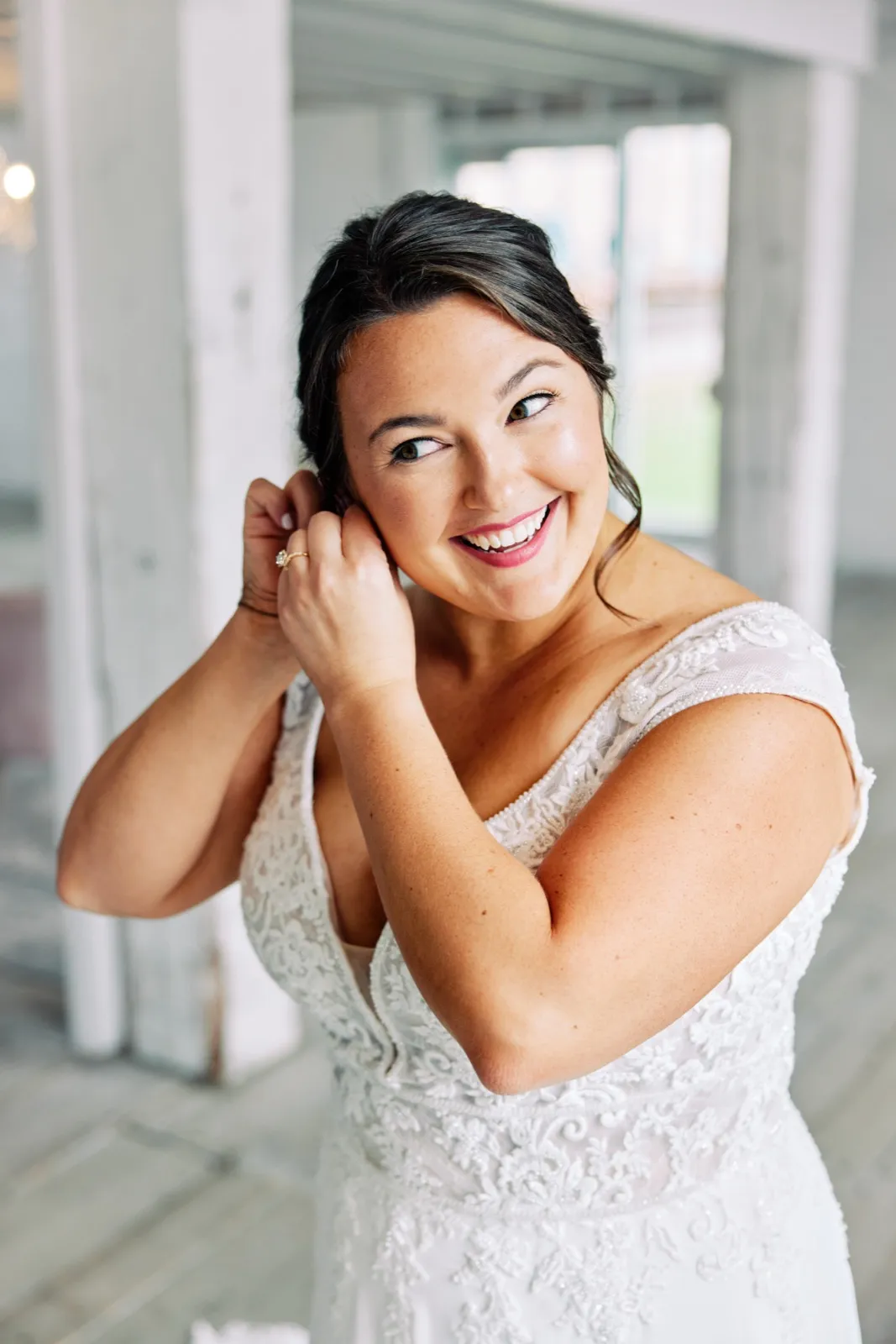 Maggie smiles toward the window while fastening an earring, the lace shoulders of her gown catching the light — Tim Larsen Photography, Brainerd Lakes MN