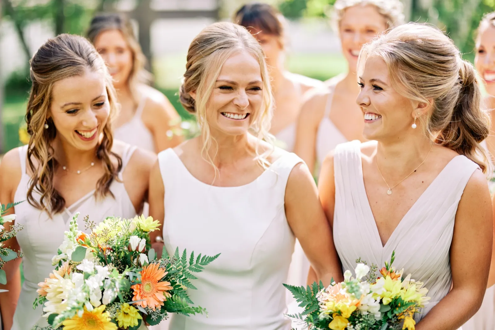 The bride laughs with her bridesmaids, all holding bouquets of greenery and orange and yellow blooms on a lawn. — Tim Larsen Photography, Brainerd Lakes MN