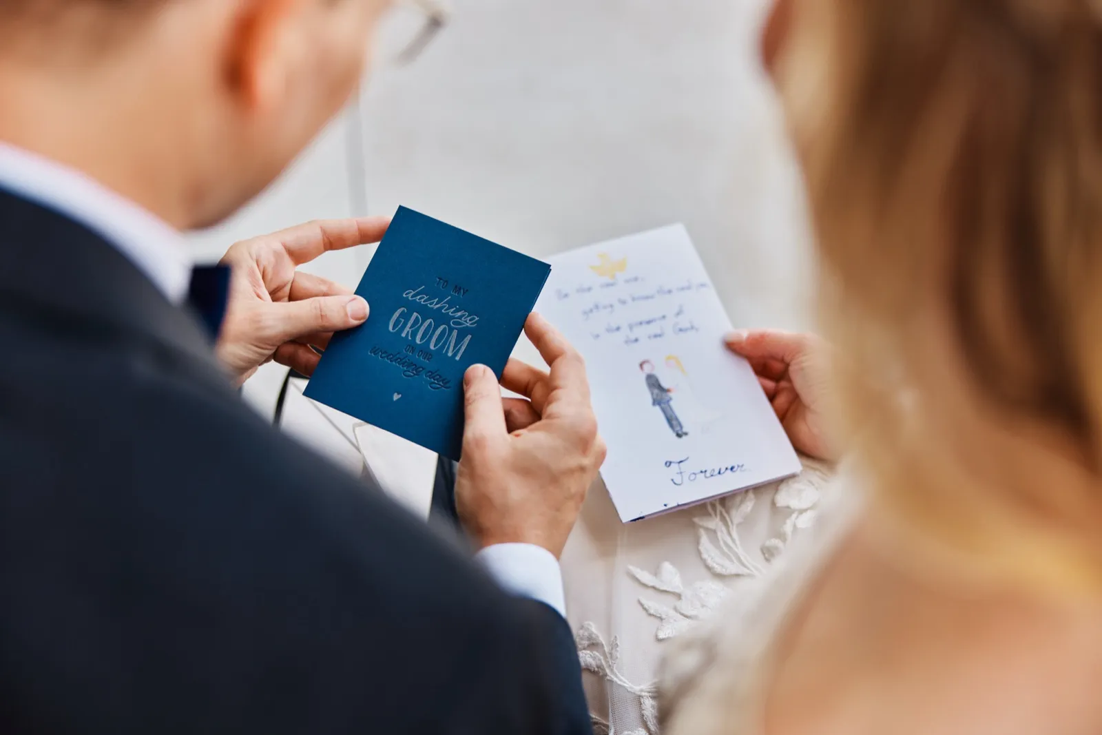 An over-the-shoulder view of the groom holding a card reading 'to my dashing groom on our wedding day' while the bride holds a hand-illustrated note ending with the word 'Forever.' — Tim Larsen Photography, Minnesota