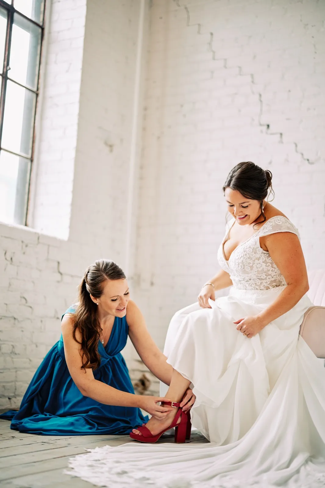 A bridesmaid in a blue dress kneels to fasten Maggie's pink heel as Maggie sits beside a tall industrial window — Tim Larsen Photography, Brainerd Lakes MN
