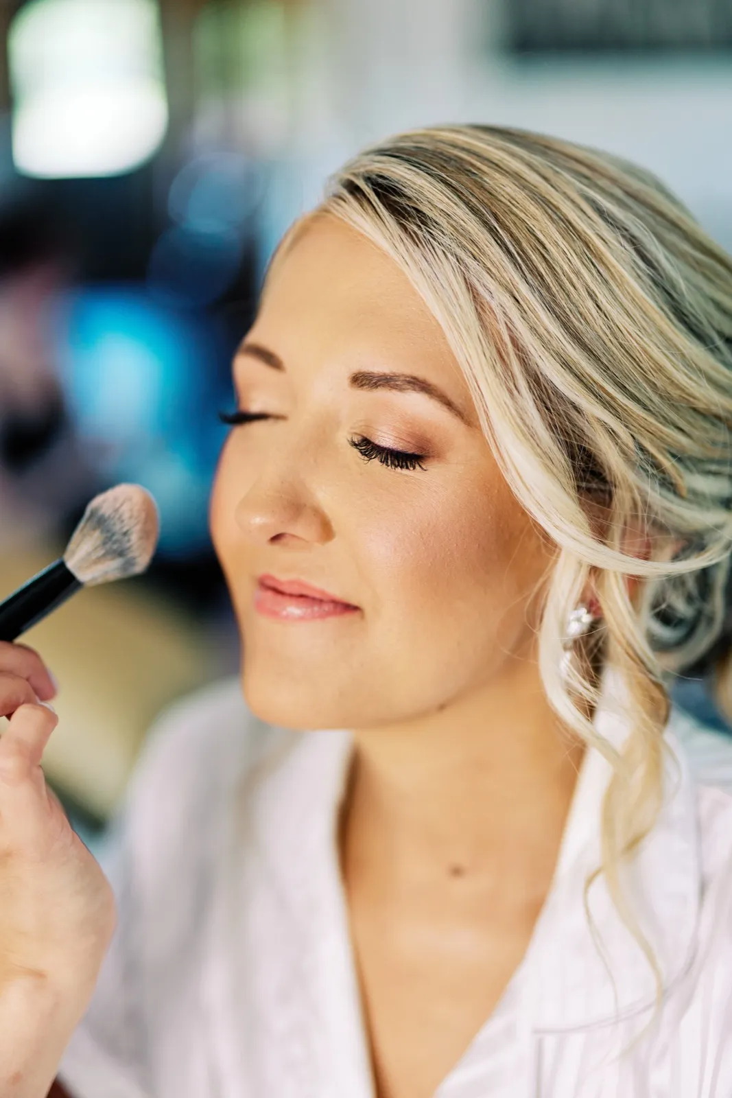 A close-up profile of the bride with eyes closed as a makeup brush is applied to her cheek in soft window light. — Tim Larsen Photography, Twin Cities MN