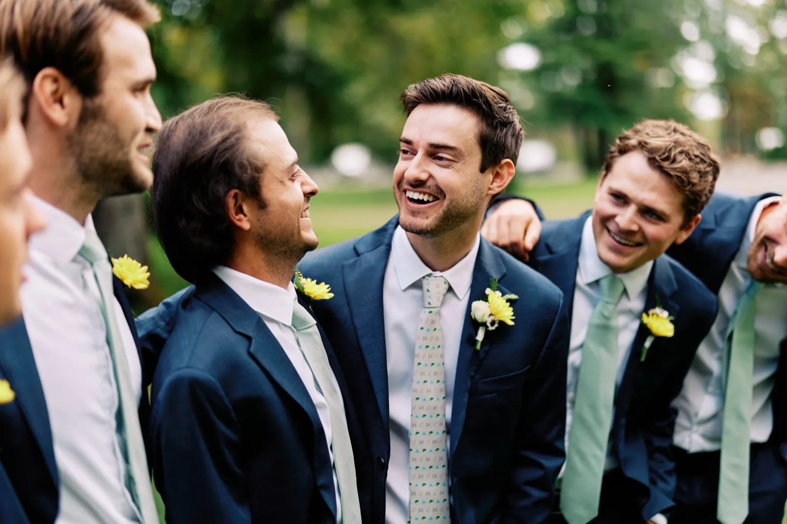 The groom laughs with his groomsmen huddled close together on a lawn, all wearing navy suits with sage ties and yellow boutonnieres. — Tim Larsen Photography, Brainerd Lakes MN