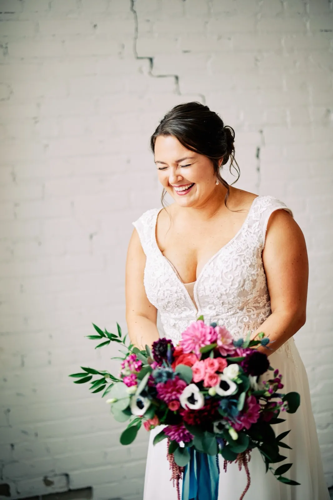Maggie laughs down at her vibrant jewel-tone bouquet of pink, magenta, and white blooms against the white brick of Le Lier — Tim Larsen Photography, Brainerd Lakes MN