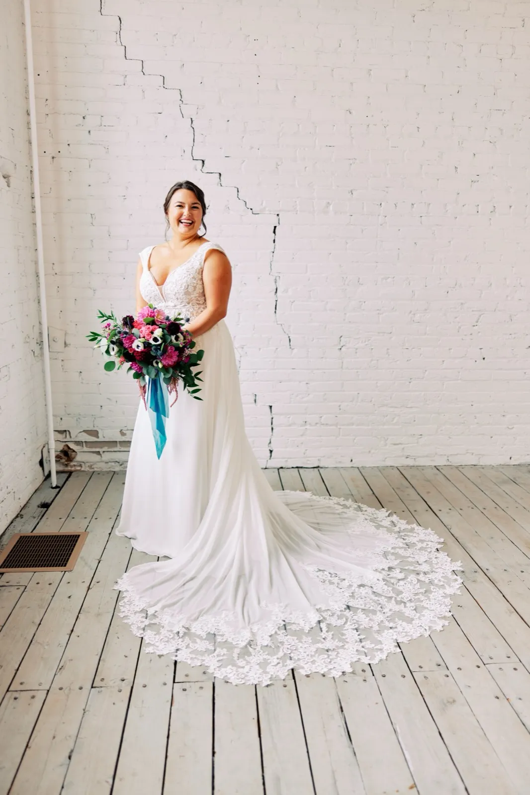 Maggie smiles with her jewel-tone bouquet and blue ribbons, her lace train fanned across the wood floor of Le Lier against the white brick — Tim Larsen Photography, Brainerd Lakes MN