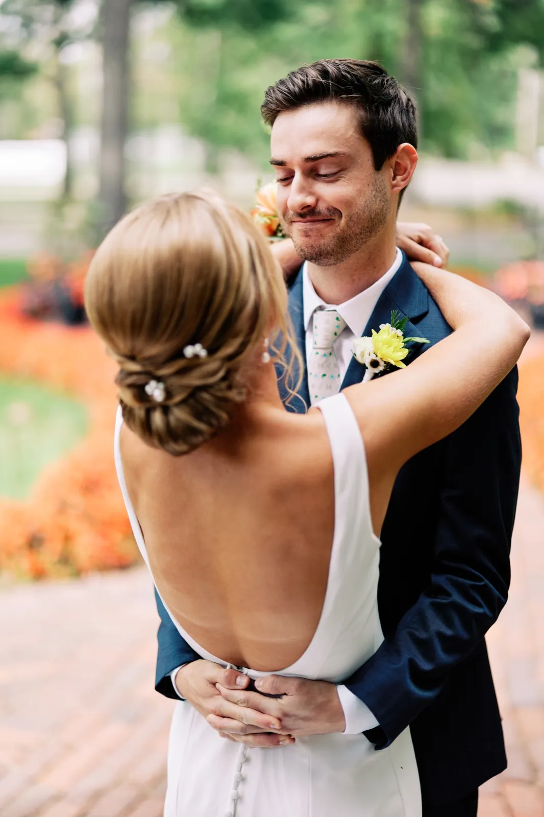 The groom rests his hands on the bride's shoulders with eyes closed and a small smile as she embraces him, her low updo and open-back dress facing the camera against a backdrop of orange autumn flowers.