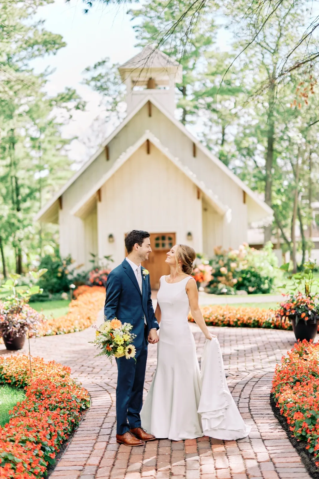 The bride and groom hold hands and gaze at each other on a brick pathway lined with orange and pink flowers, with the white chapel at Grand View Lodge rising behind them. — Tim Larsen Photography, Brainerd Lakes MN