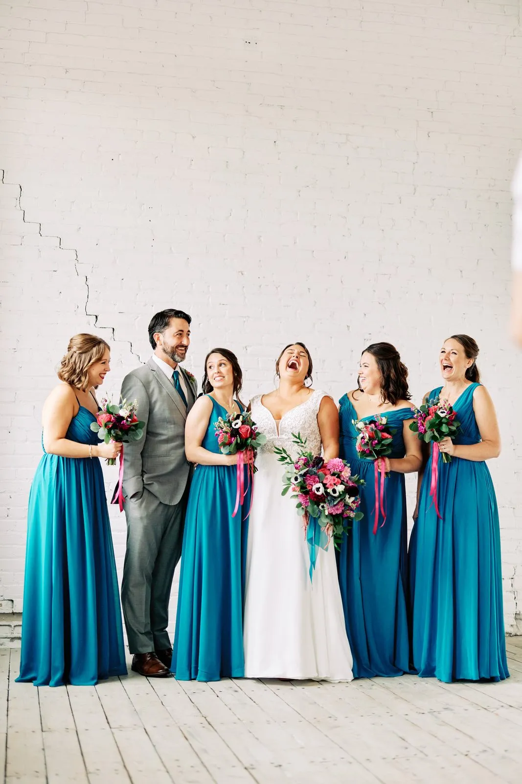 Maggie throws her head back laughing with her wedding party in blue dresses and a gray suit against the whitewashed brick wall — Tim Larsen Photography, Brainerd Lakes MN