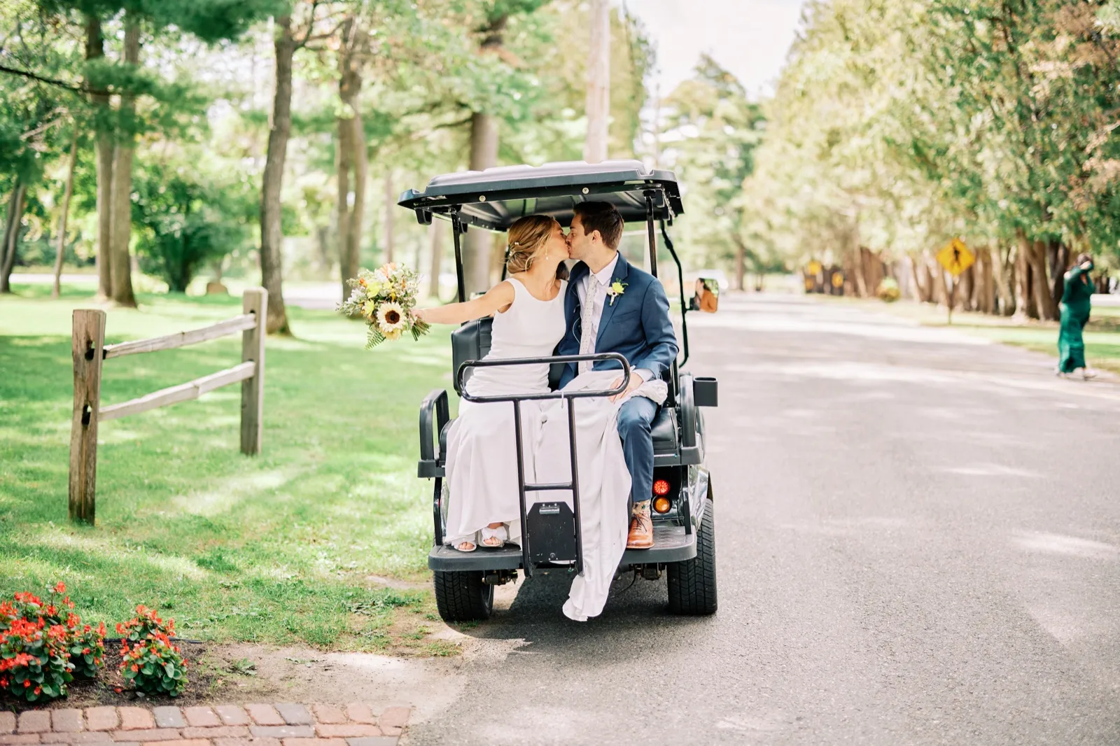 The bride and groom kiss in the back of a golf cart on a tree-lined resort road, the bride holding a sunflower bouquet. — Tim Larsen Photography, Brainerd Lakes MN