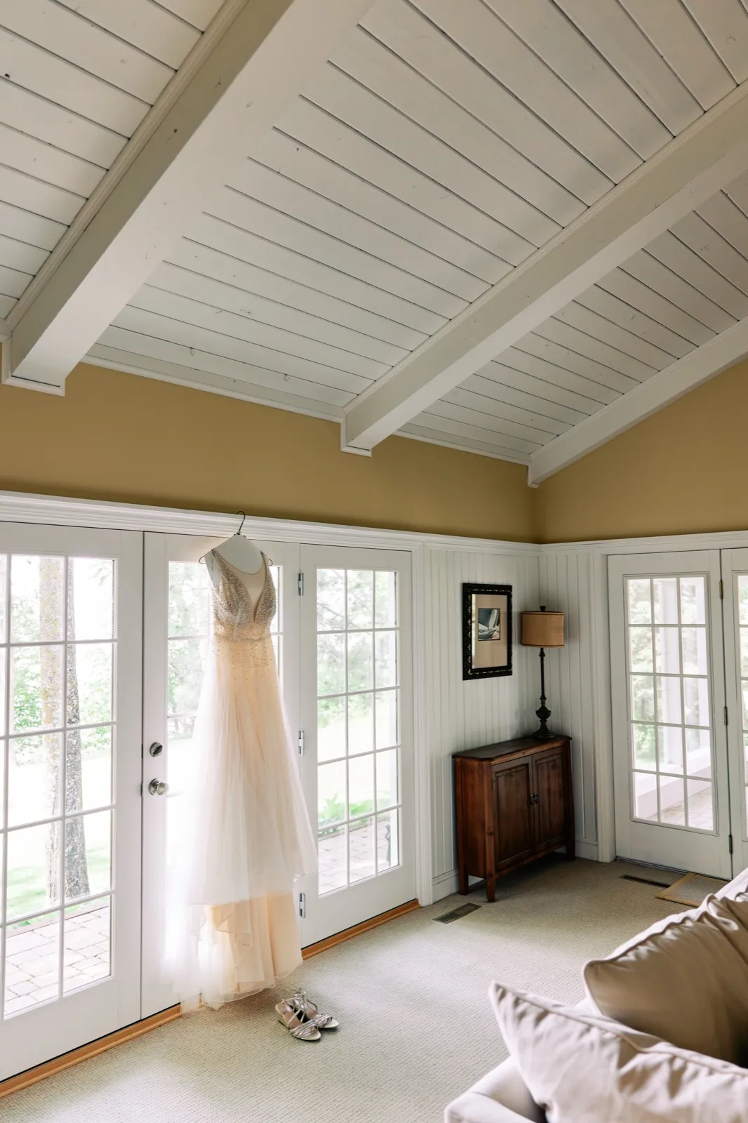 A blush wedding gown hangs from the trim of a French door in a vaulted-ceiling sunroom, with the bride's strappy shoes placed on the carpet below. — Tim Larsen Photography, Brainerd Lakes MN