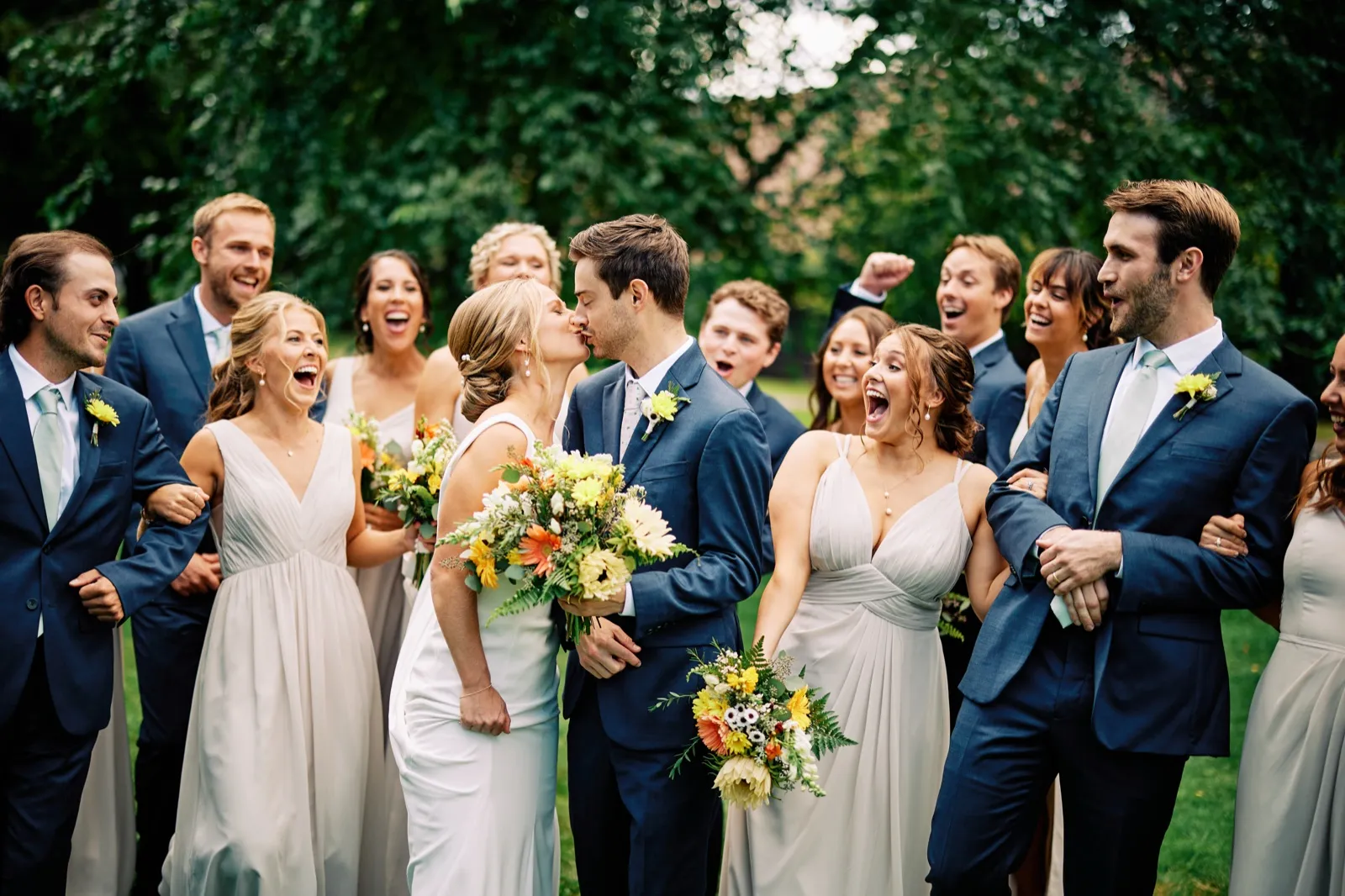 The bride and groom kiss at the center of their bridal party as friends around them cheer and laugh in surprise on a green lawn. — Tim Larsen Photography, Brainerd Lakes MN
