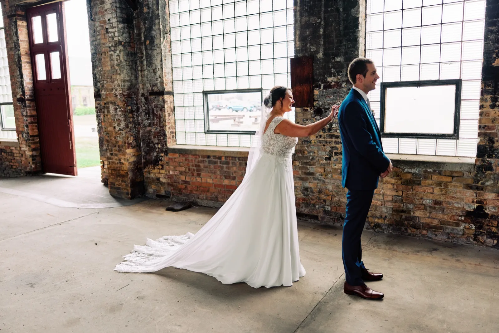 Maggie reaches to tap Craig's shoulder in a sunlit warehouse with exposed brick walls and tall grid windows — Tim Larsen Photography, Brainerd Lakes MN