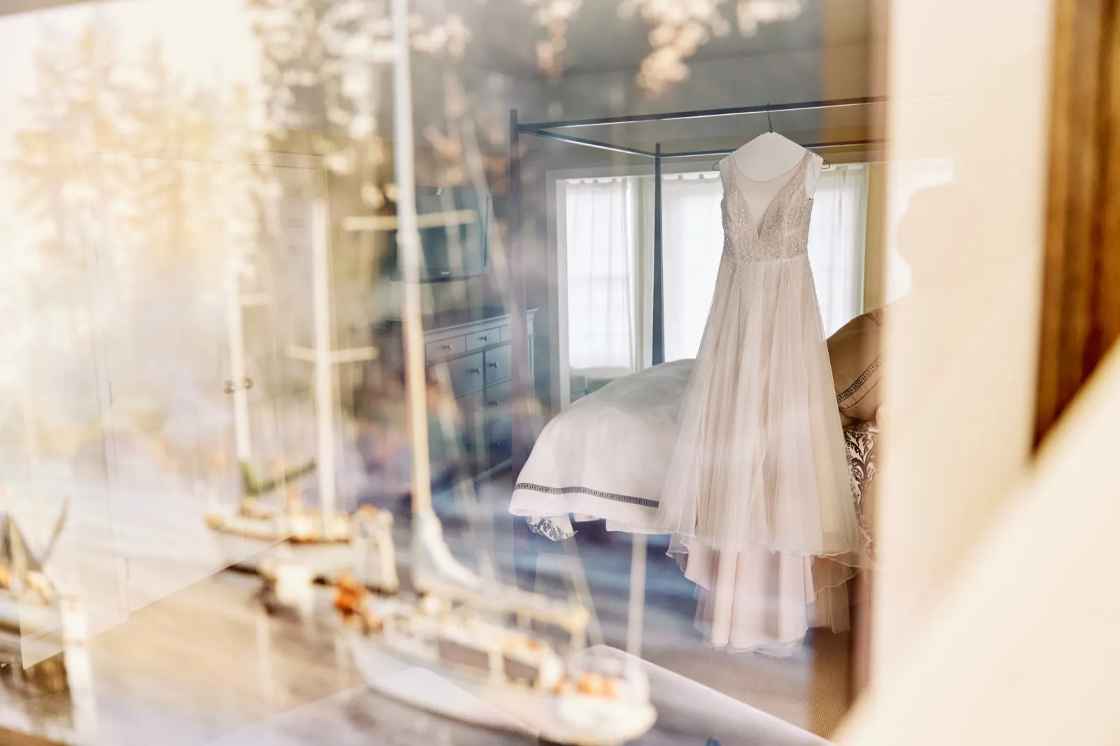 A beaded wedding gown hangs from a four-poster bed in a sunlit bedroom, photographed through a glass case containing model sailboats that reflects trees outside. — Tim Larsen Photography, Brainerd Lakes MN