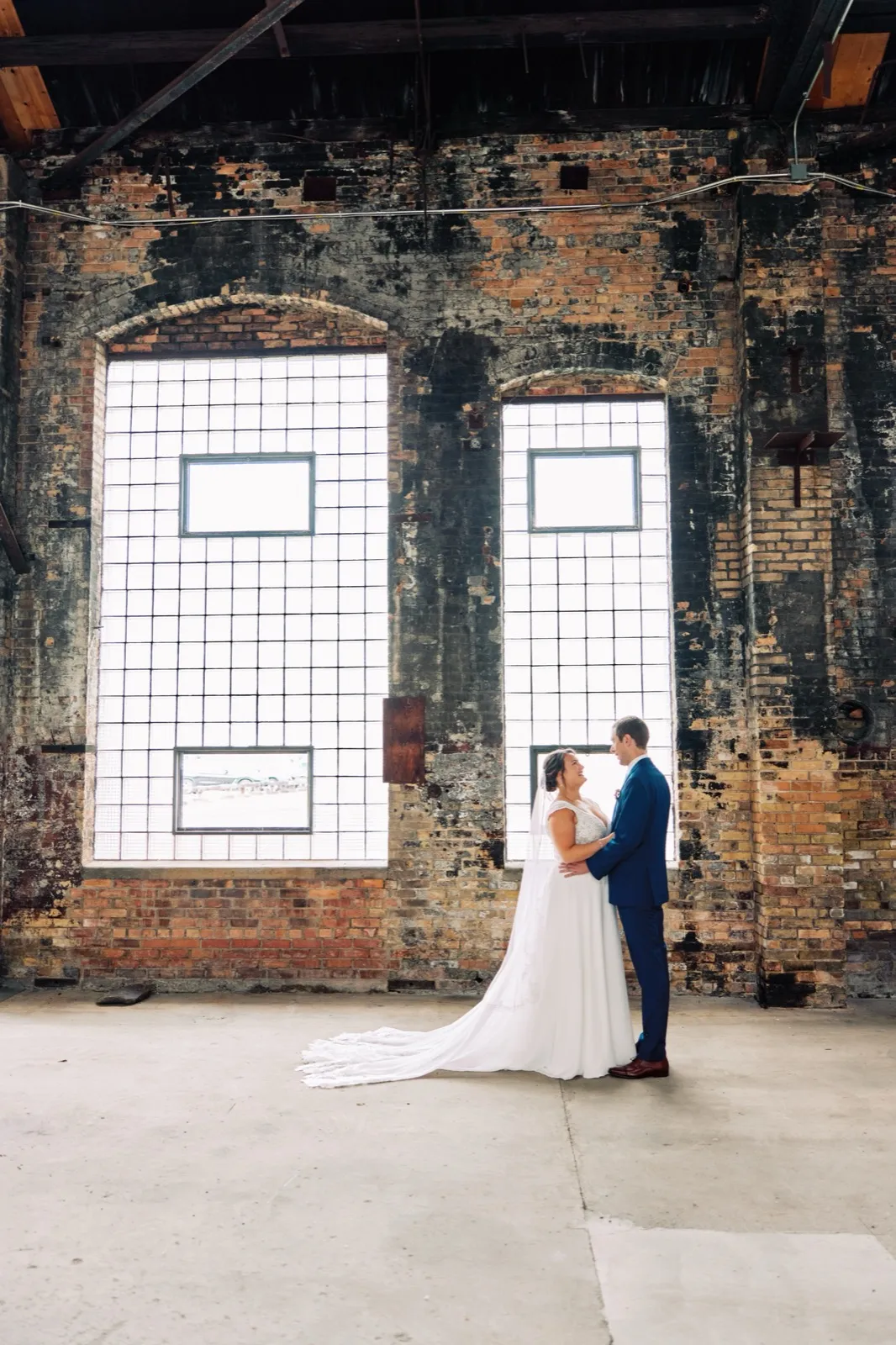Maggie and Craig embrace beneath two towering arched grid windows in a weathered brick warehouse interior, her long train trailing across the concrete — Tim Larsen Photography, Brainerd Lakes MN