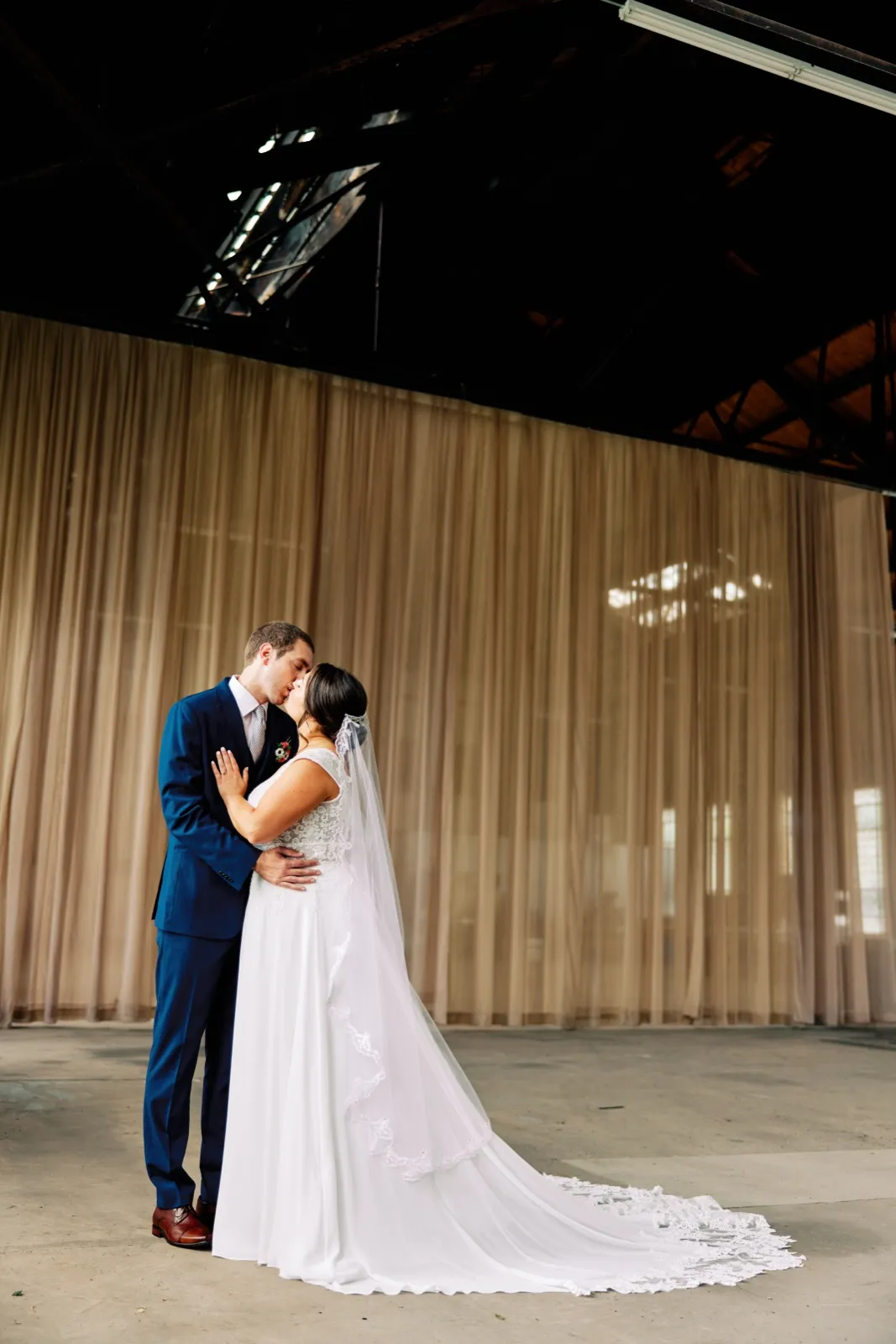 Maggie and Craig kiss beneath exposed steel trusses in a tall draped hall, her veil and lace train spread across the concrete — Tim Larsen Photography, Brainerd Lakes MN