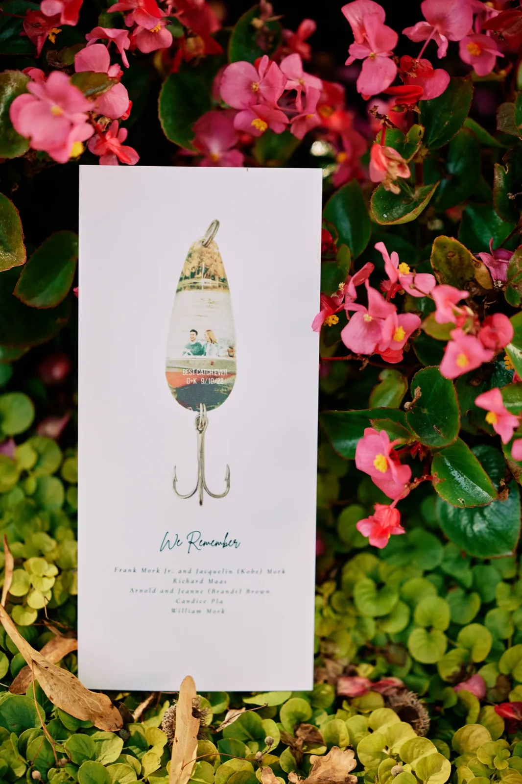A wedding program shaped like a fishing lure with the words 'Best Catch Ever' and a 'We Remember' memorial list rests among bright pink begonias and green foliage. — Tim Larsen Photography, Brainerd Lakes MN