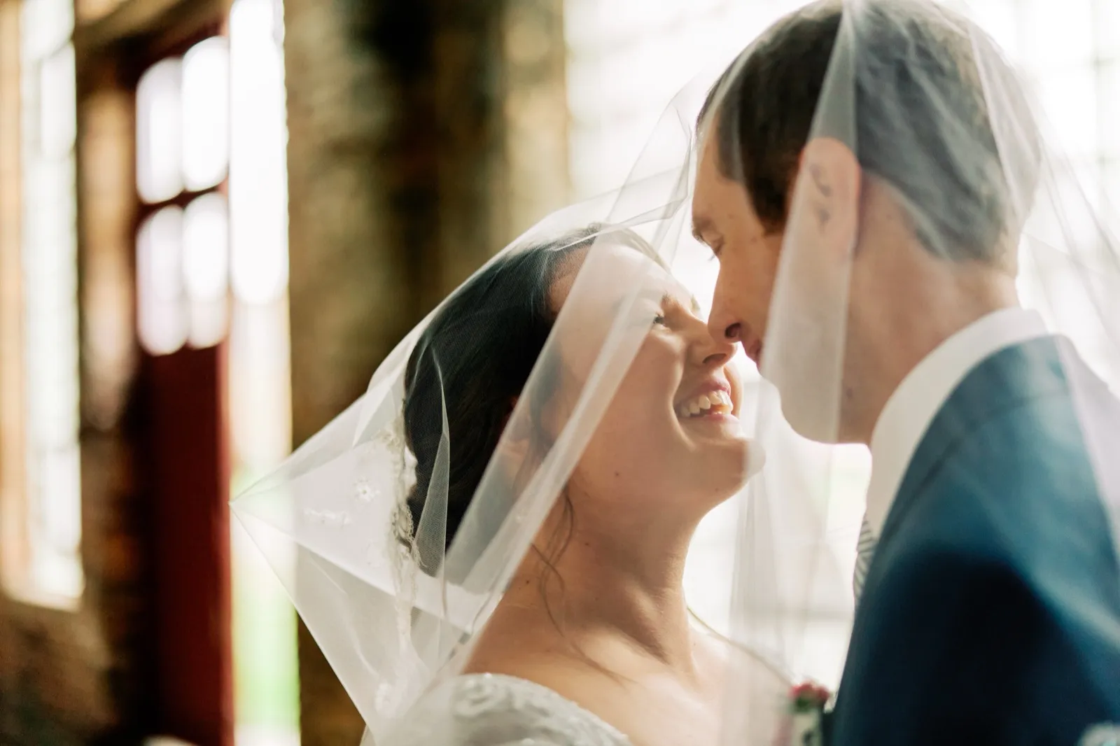 Maggie laughs with her head tilted up toward Craig as her veil drapes over both of them in warm window light — Tim Larsen Photography, Brainerd Lakes MN