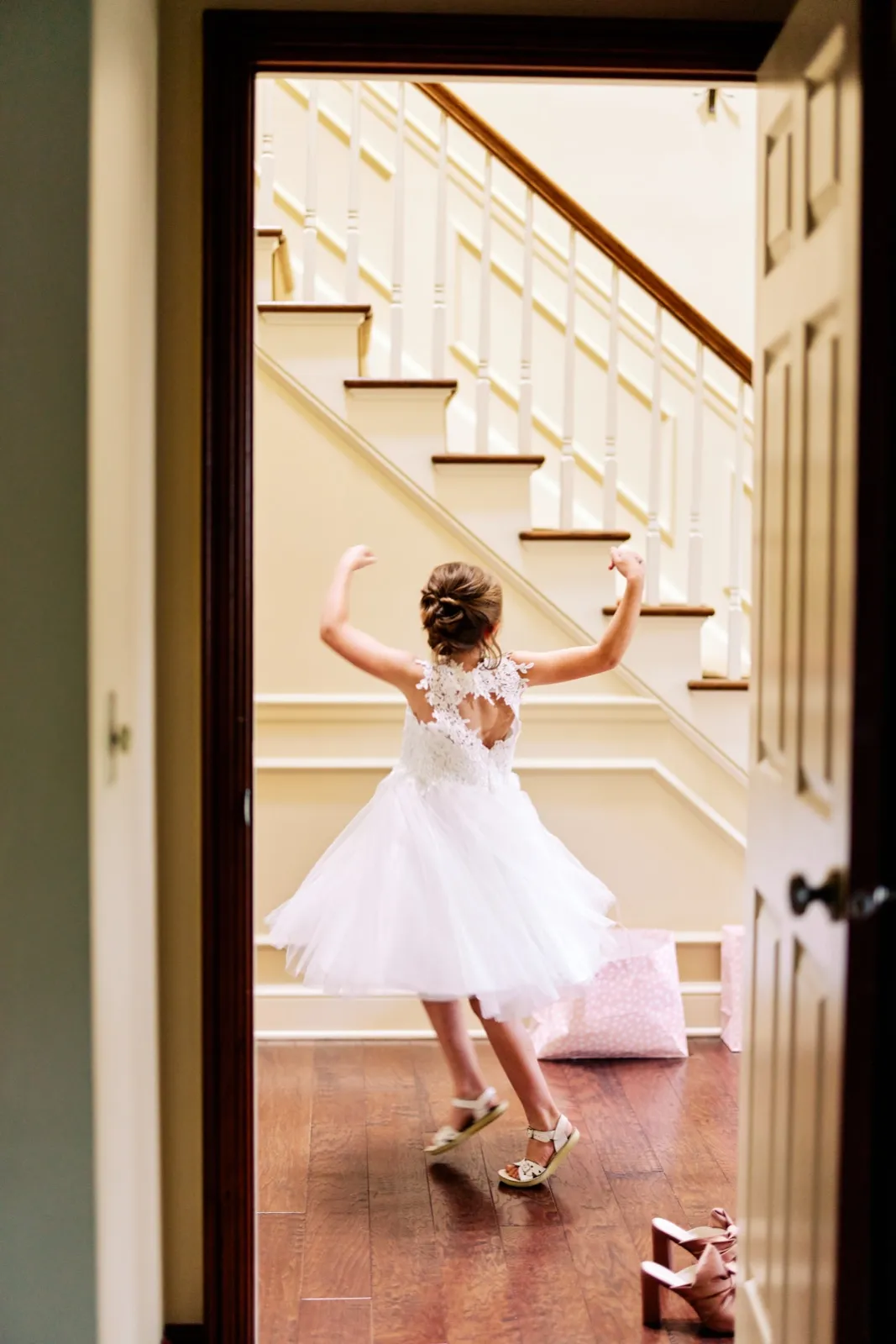A flower girl in a white tulle dress twirls with arms raised at the foot of a staircase, viewed through an open doorway. — Tim Larsen Photography, Brainerd Lakes MN