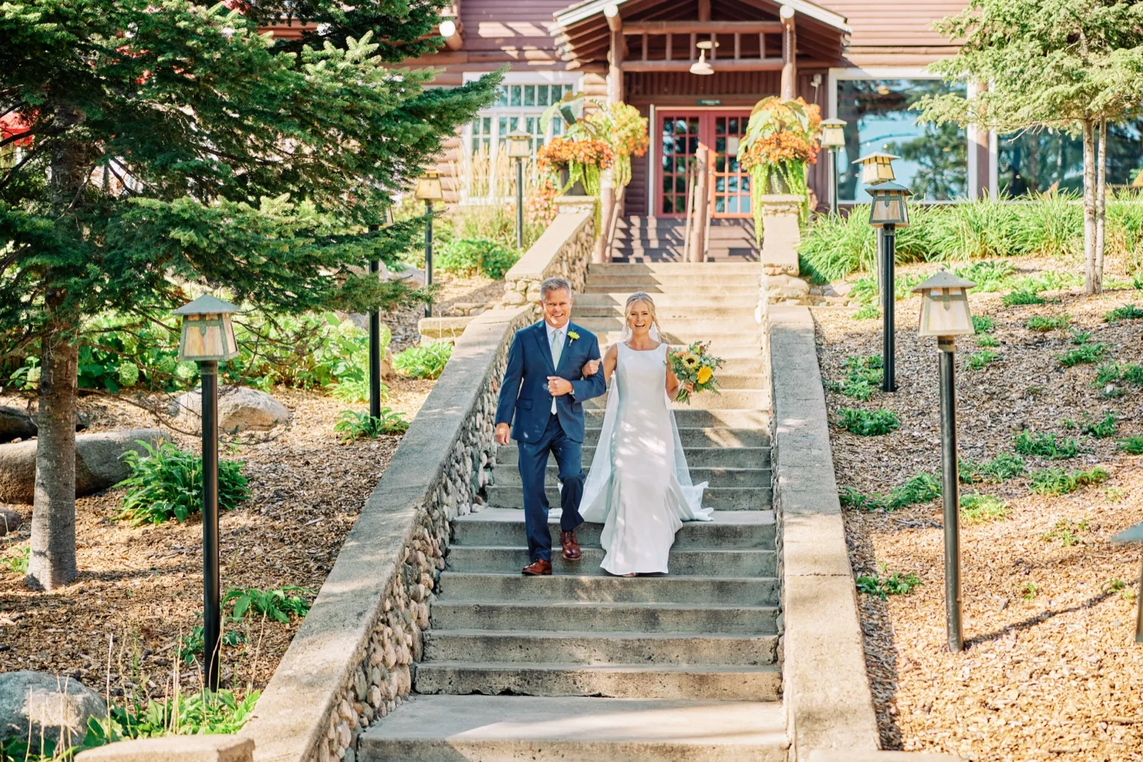 The bride and groom walk down a wide stone stairway flanked by craftsman lanterns and evergreens, with the timber lodge entrance behind them. — Tim Larsen Photography, Brainerd Lakes MN