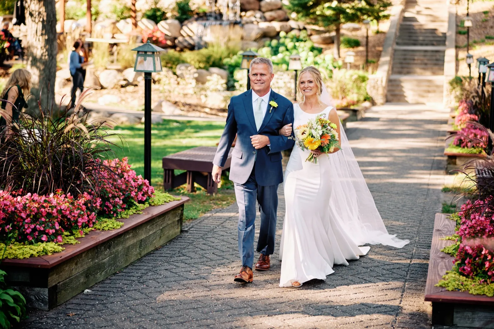 The bride walks down a stone aisle on her father's arm, holding a bright sunflower bouquet, flanked by pink flower beds at Grand View Lodge. — Tim Larsen Photography, Brainerd Lakes MN
